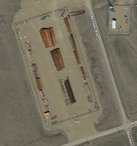 Screenshot of a rectangular fenced dirt lot as seen from google maps somewhere in Montana. The lot contains a few piles of wood, and the the way the dirt extends beyond the fencing and interacts with the road is highly reminiscent of Cities Skylines